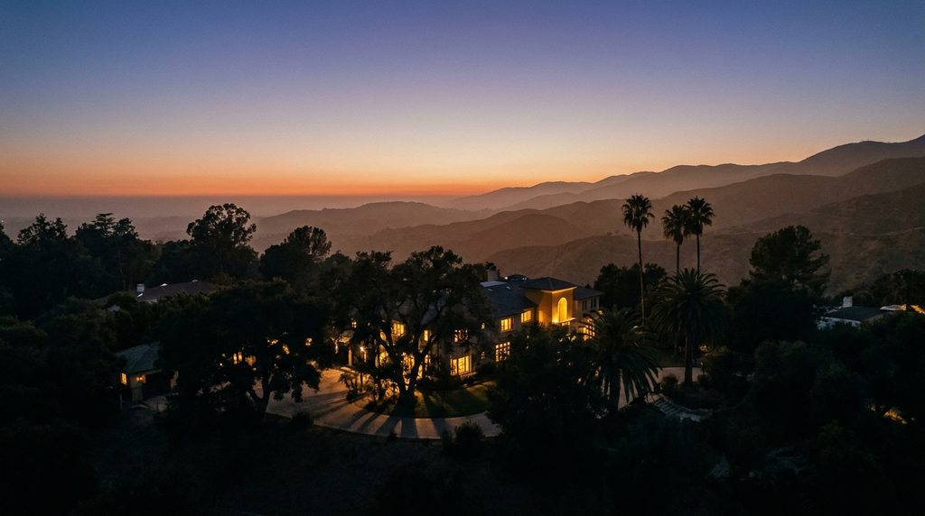 An aerial view at dusk over a hillside neighbourhood, lit windows among oaks and palms with mountains behind.
